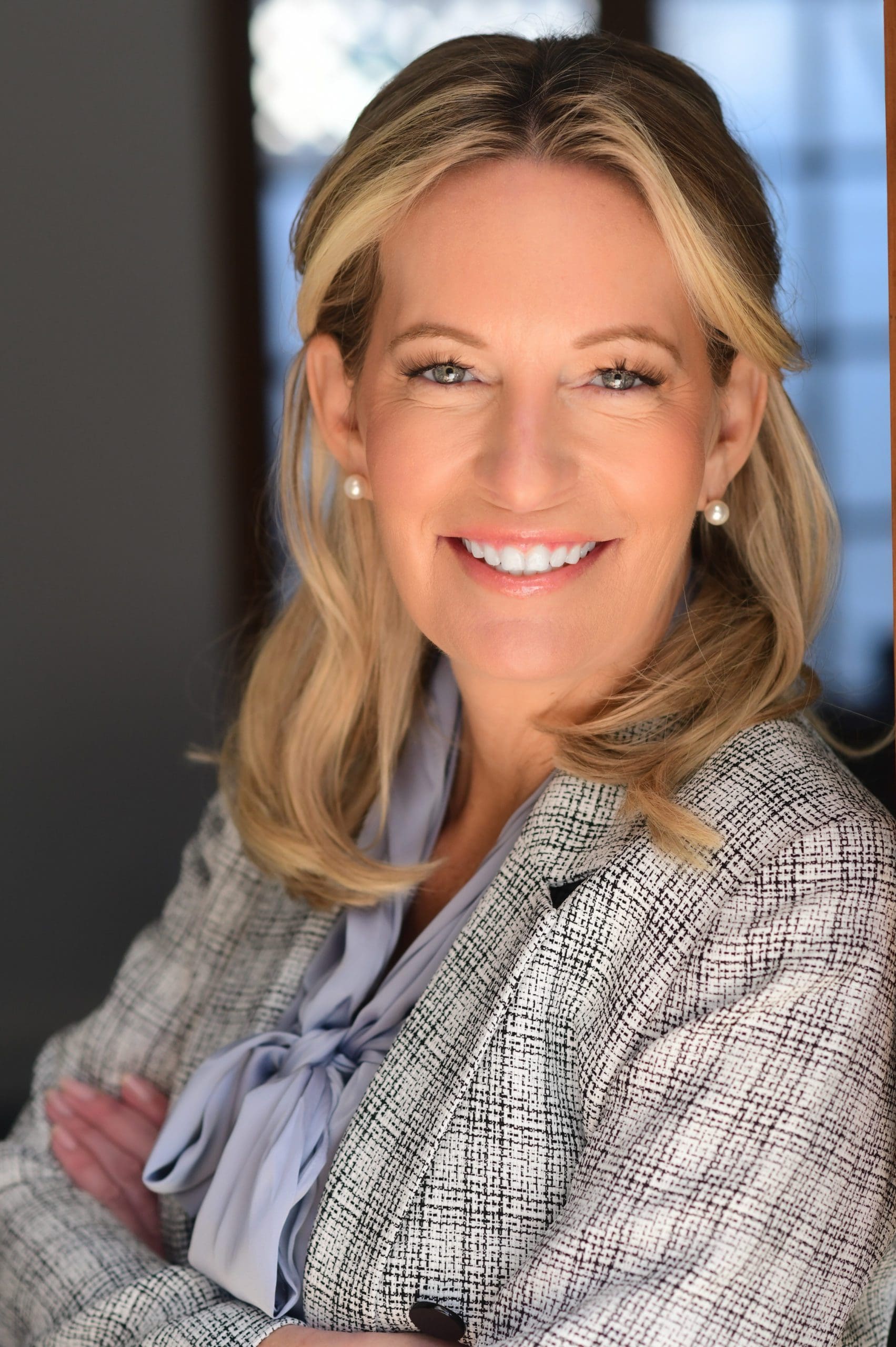 Attorney Kelly Hyman, with a warm smile, presents a professional demeanor at The Hyman Law Firm P.A., located in West Palm Beach, Florida. Her blonde hair and polished appearance, featuring a stylish tweed blazer and light blue blouse, complement the modern office backdrop with expansive windows that hint at the West Palm Beach urban landscape.