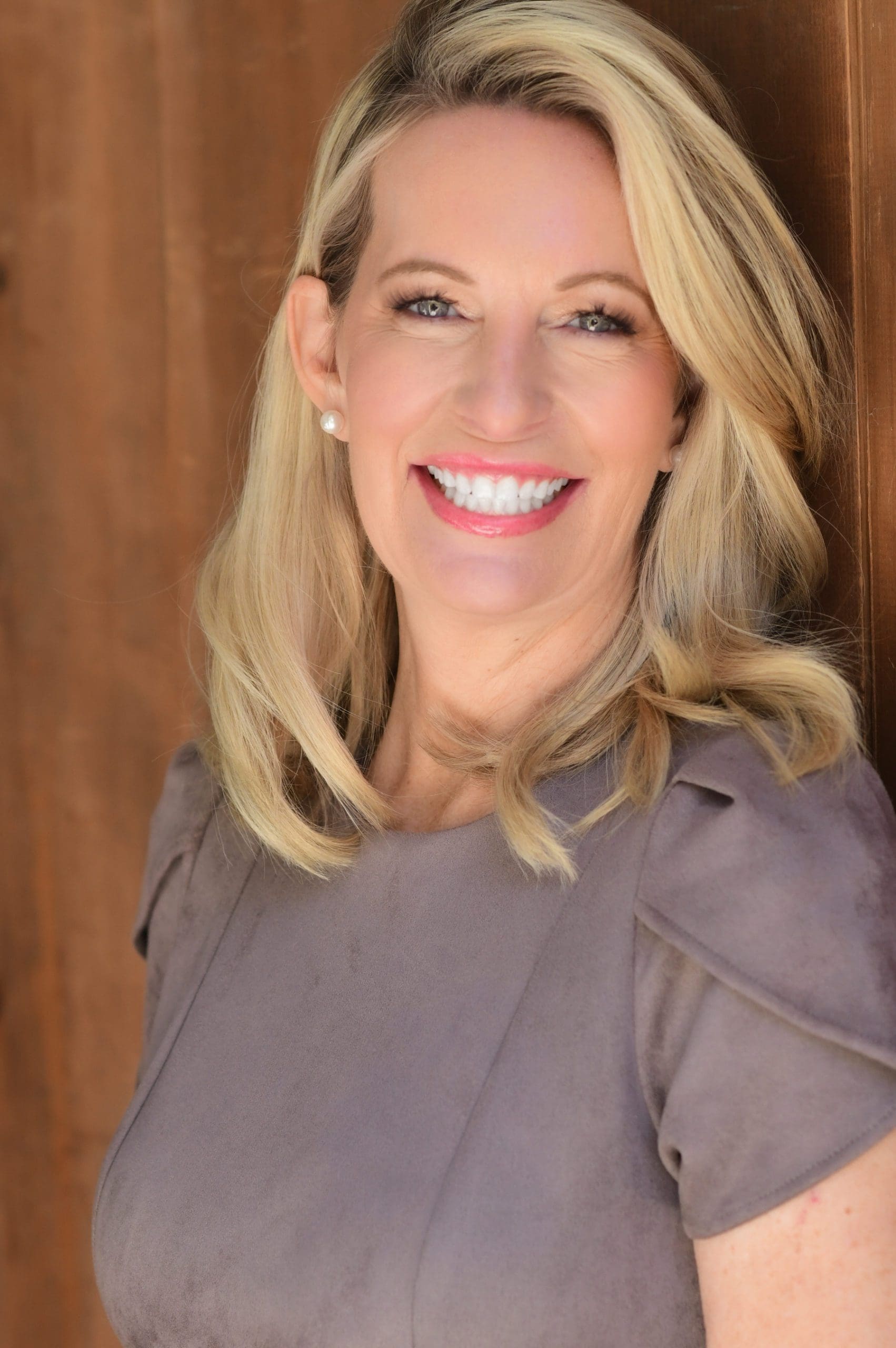Portrait of Kelly Hyman, attorney at The Hyman Law Firm P.A., featuring her engaging smile against a warm wooden background. She is dressed in a professional grey outfit with pearl earrings, capturing a blend of approachability and professionalism.