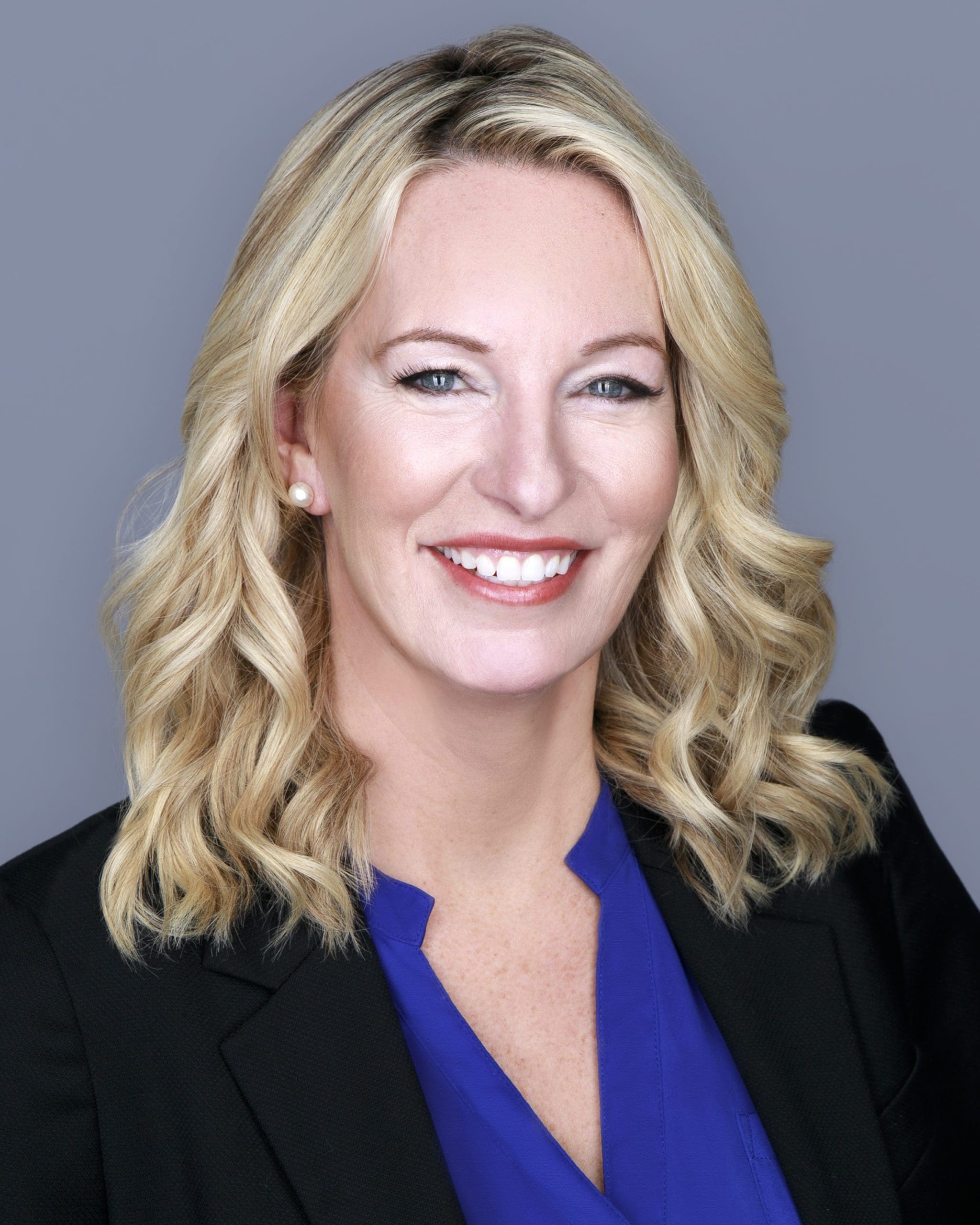 Headshot of Kelly Hyman, attorney at The Hyman Law Firm P.A., with a beaming smile. She has blonde, wavy hair and is wearing professional attire including a black blazer and a blue top, complemented by pearl earrings. The background is a neutral gray, highlighting her features and professional appearance."